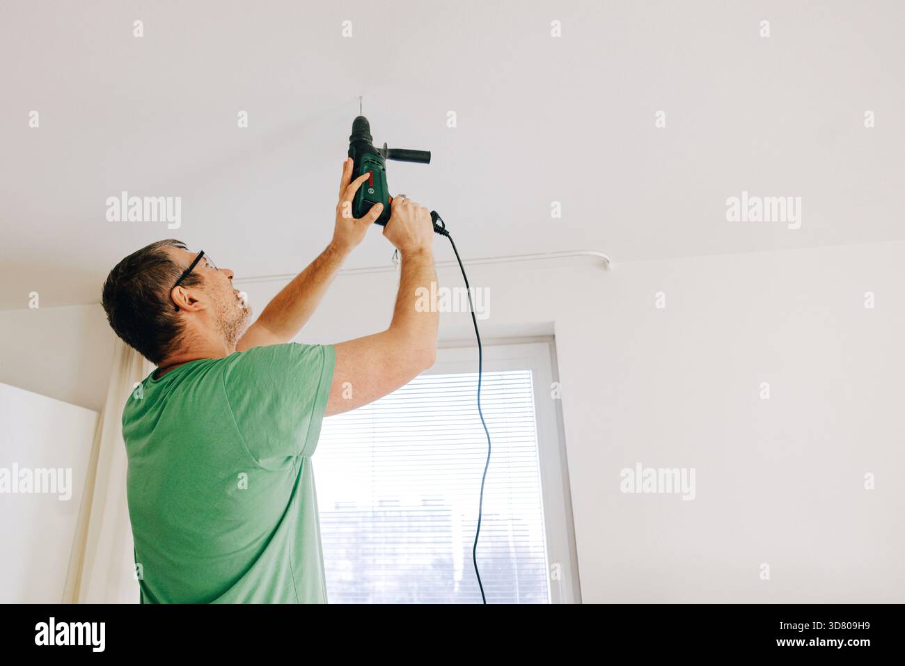 Man using electric drill to make hole in white ceiling at home. Stock Photo