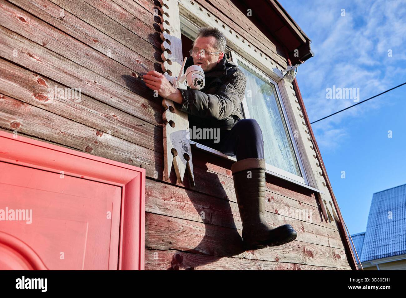 Pulling surveillance camera cable through drilled passage in wall, installer sits on window frame and feeds wire toward exterior mounting point. Stock Photo