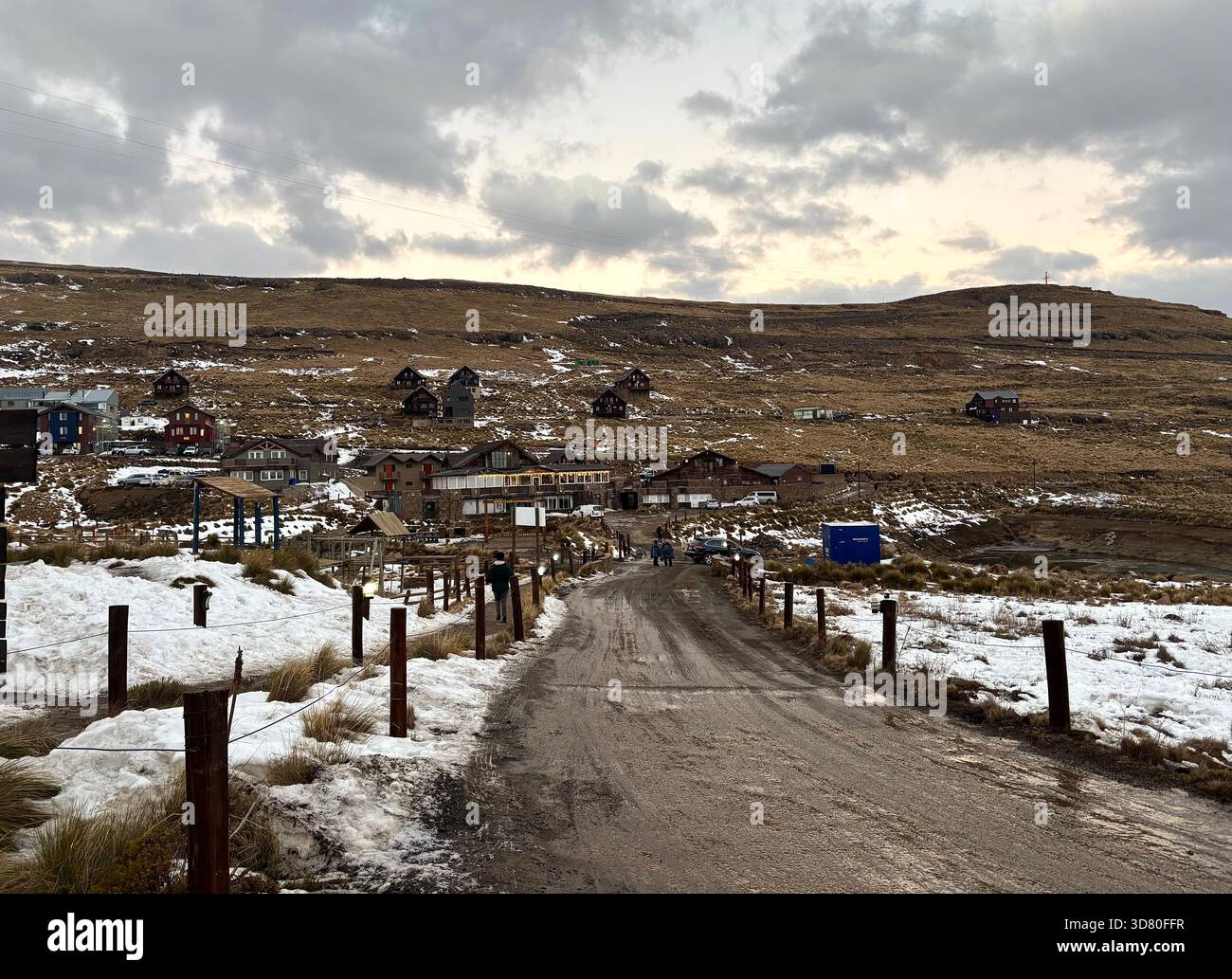 Snow-covered mountain village at Afriski Resort in Lesotho during winter, with scattered house, muddy road, and dramatic cloudy sky - Smartphone Captured Stock Image
