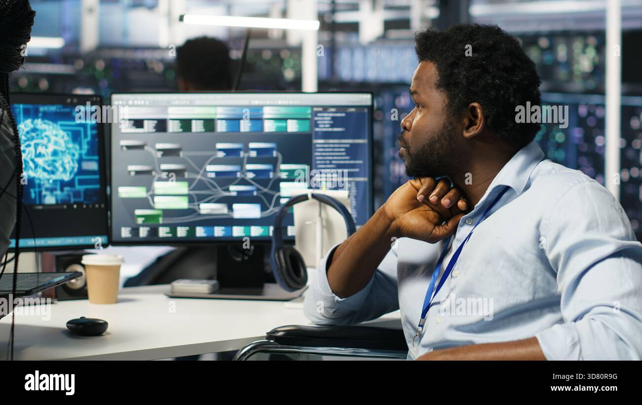 Data center engineer in wheelchair and colleague oversee automated tasks using node tree programming tool. Inclusive server farm team adjusts system settings based on AI analytics, camera B Stock Photo