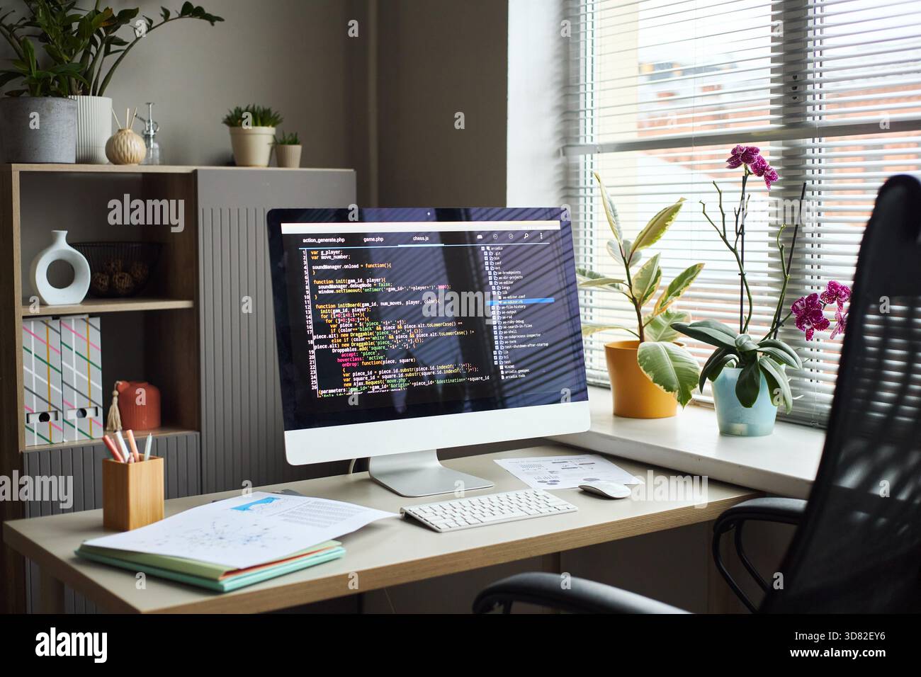 Young Asian man working at desk using desktop computer with programming code on screen, surrounded by office plants and documents, modern office environment visible through window blinds Stock Photo