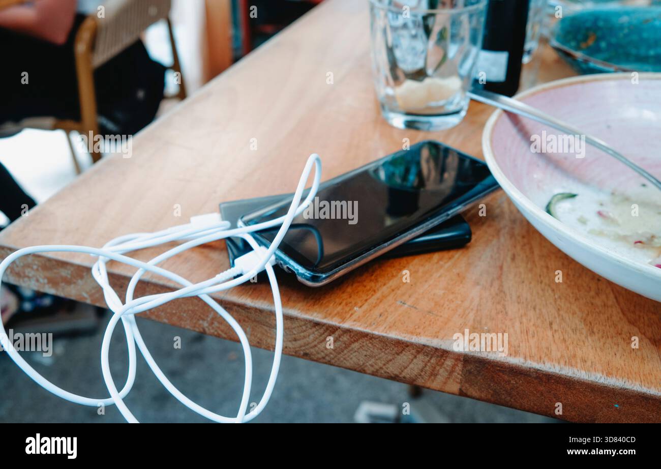 a smartphone with its white charging cable connected to a power bank rests on a wooden restaurant table, beside an empty glass and a bowl with food re Stock Photo