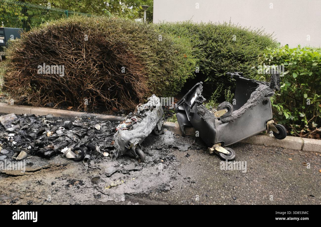 A black wheeled trash receptacle lies broken and charred on the pavement. A pile of burnt trash lies nearby, scattered on the ground near green bushes - Smartphone Captured Stock Image