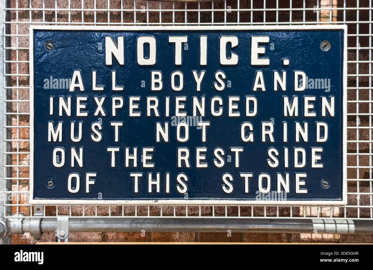 Metal sign with warning text restricting boys and inexperienced men from grinding on one side of a stone, mounted on a wire mesh fence - Smartphone Captured Stock Image