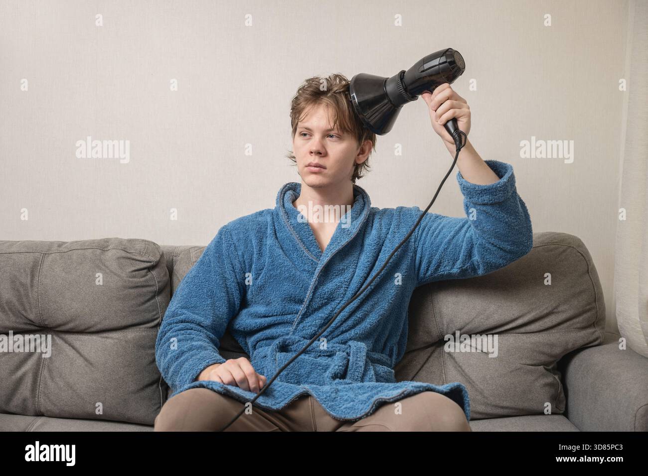 Young caucasian man in blue bathrobe using black hairdryer to dry his hair while sitting on sofa. Mens Home Self-Care Routine Stock Photo
