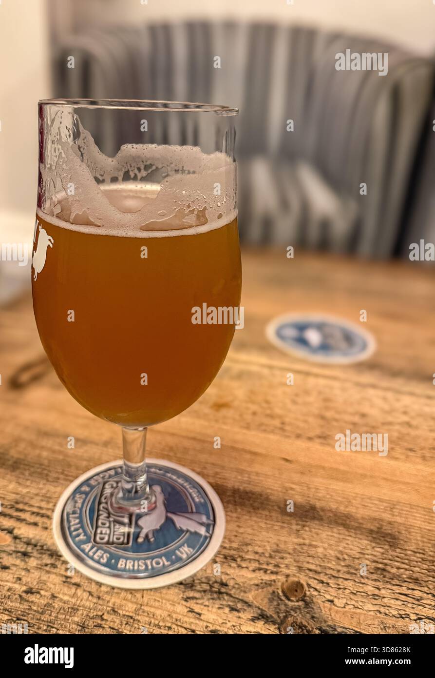 Close-up of a pint of craft beer on a rustic wooden table with a blurred striped armchair in the background - Smartphone Captured Stock Image