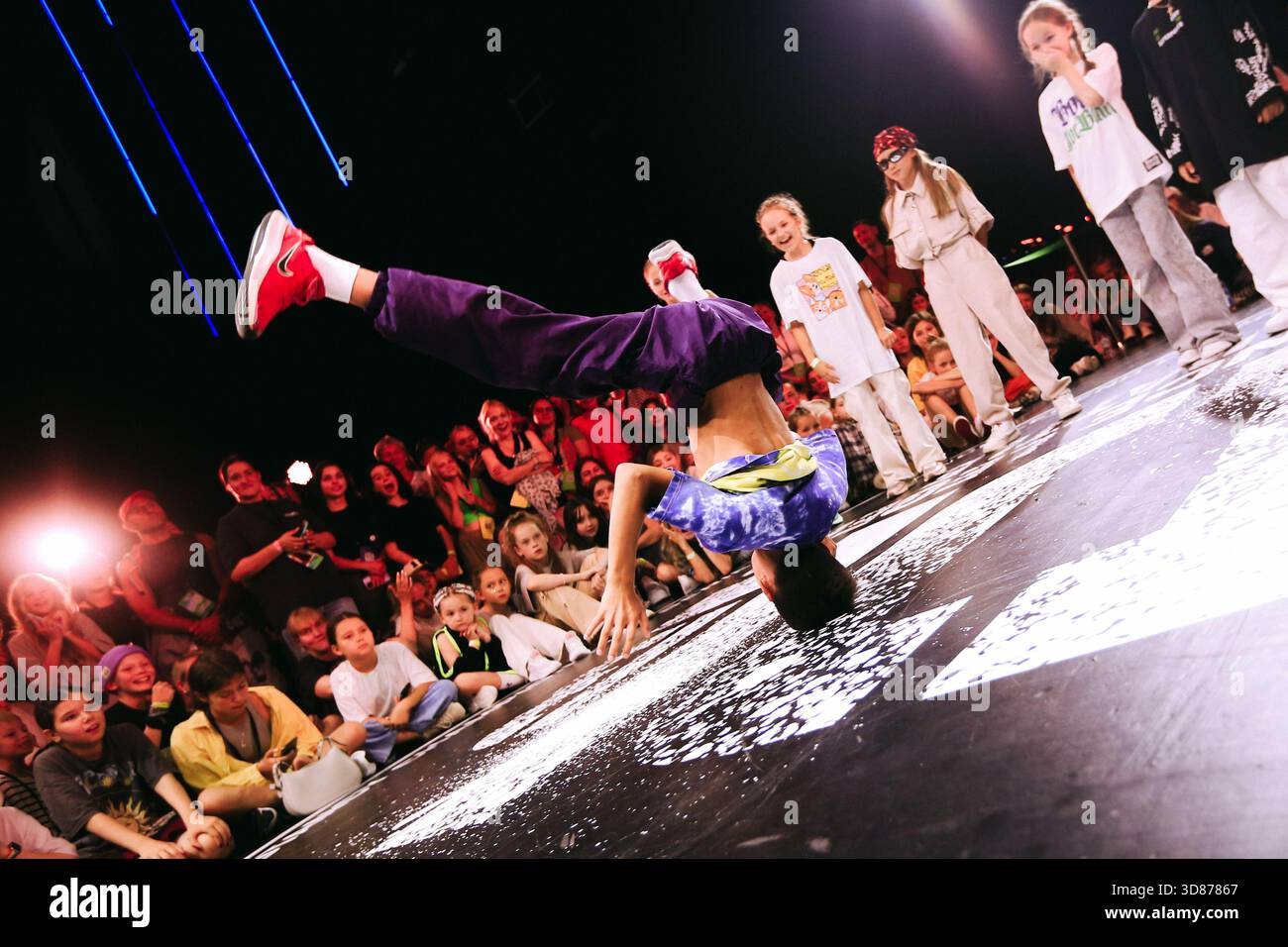 Street dancer in Blue cap breaker handstand freeze circle audience cheering under neon lights, paint-splattered floor, victorious pose after round win Stock Photo