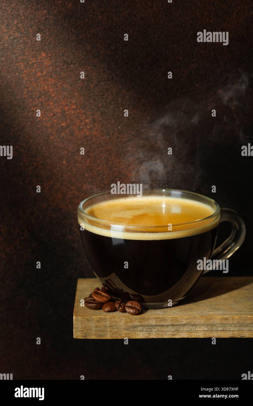 Ray of light falls on transparent cup of coffee with beans scattered around it against brown background Stock Photo