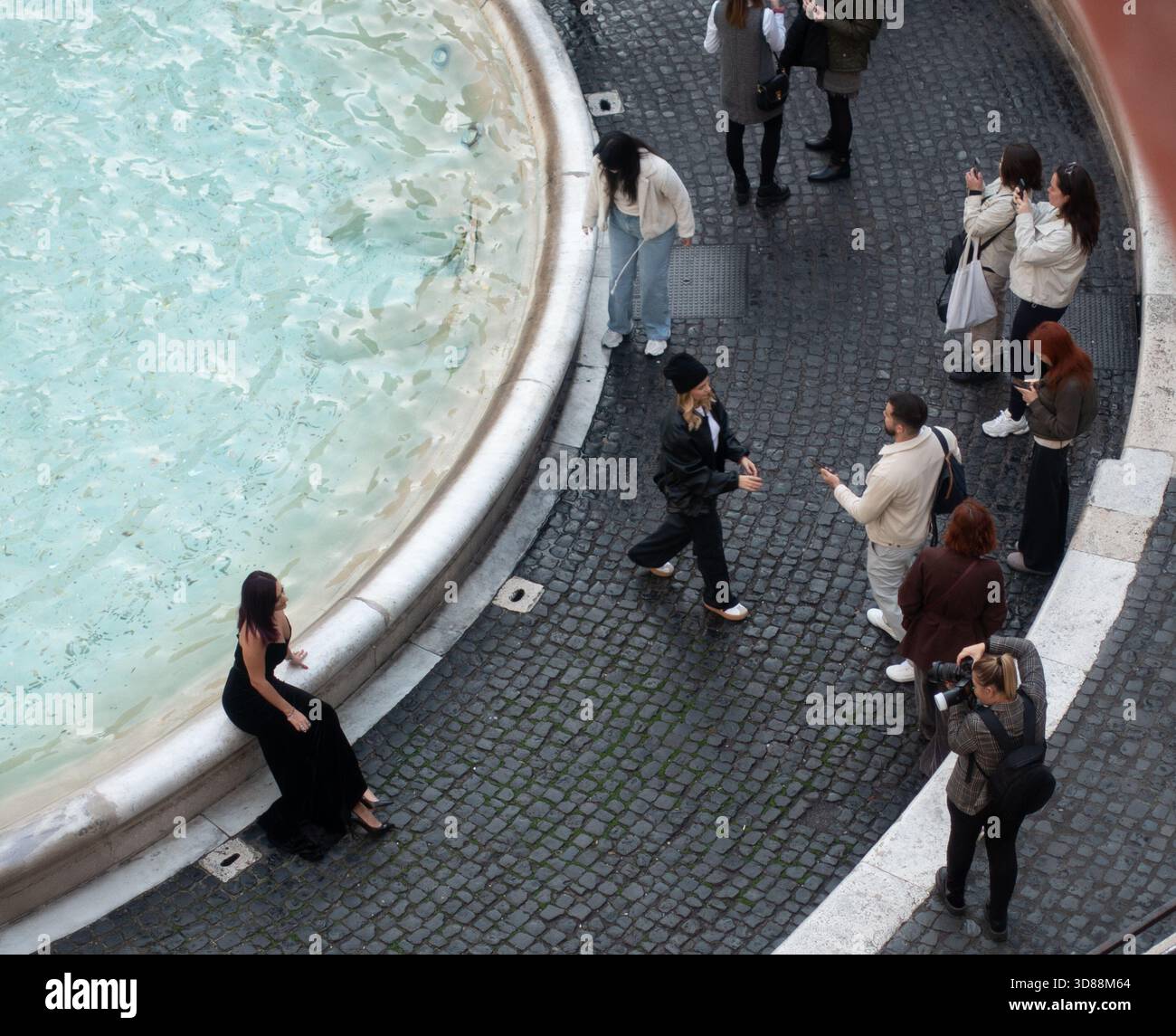 A woman in a black dress sits on the edge of the Trevi fountain while a photographer takes pictures of her. Other people are standing around watching Stock Photo