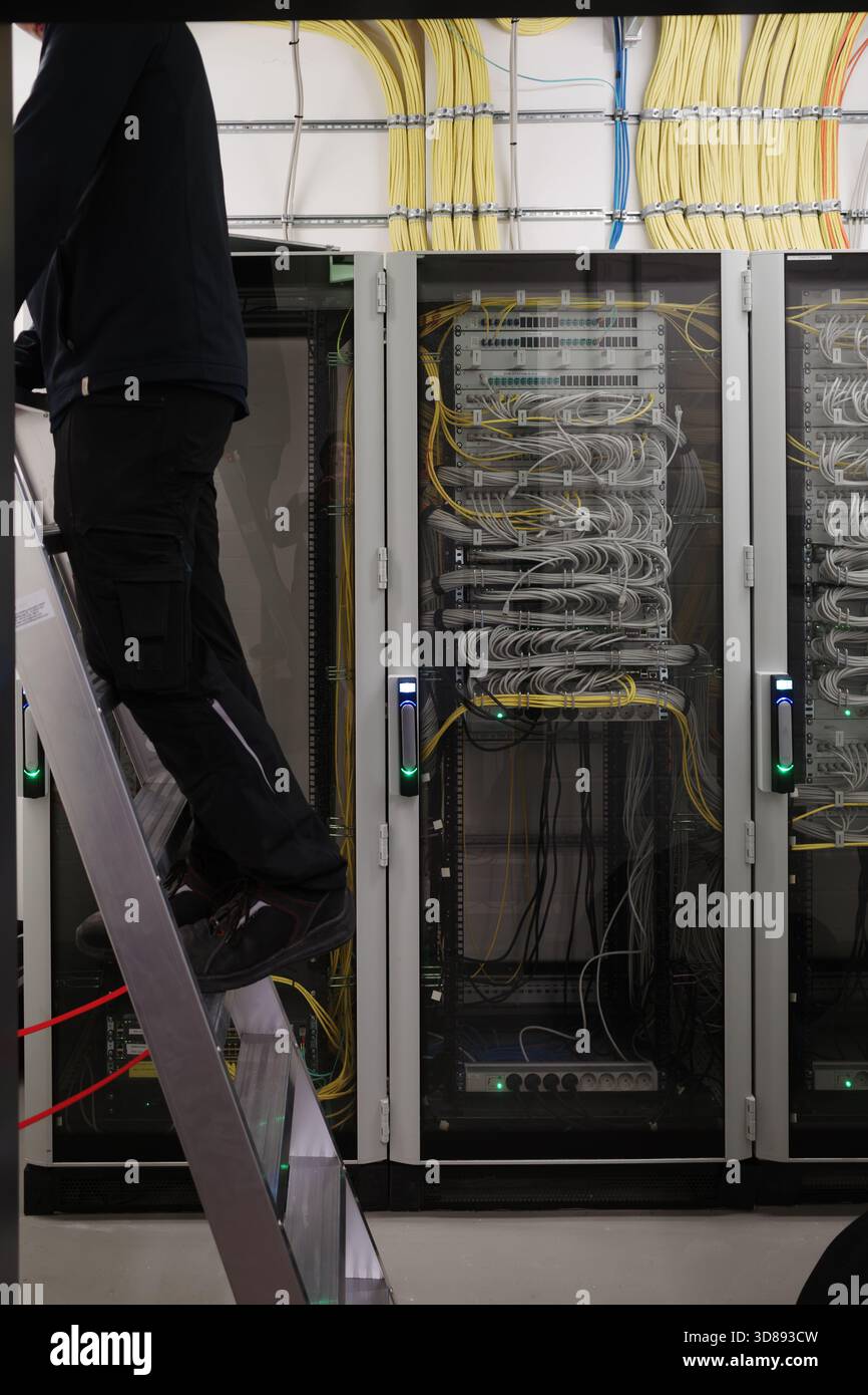 Detailed view of a modern server room with network cabinets full of organized fiber-optic and Ethernet cables. A technician standing on a ladder works Stock Photo