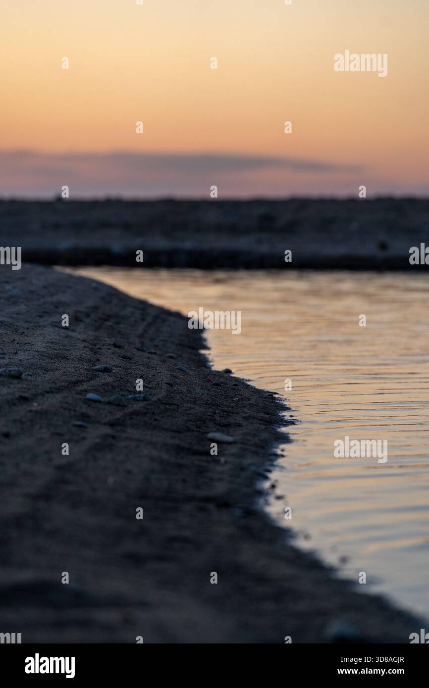 coastal morning scenery, gentle tide meets shores at sunrise under orange sky, beautiful dawn over coastline showcasing sandy curves and pebbled beach Stock Photo