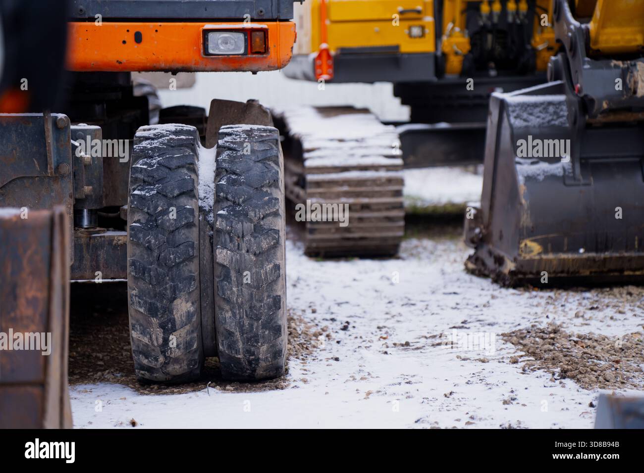 Dual construction vehicle wheels standing on snowy ground with tracked excavators and metal buckets visible in the background during winter worksite o Stock Photo
