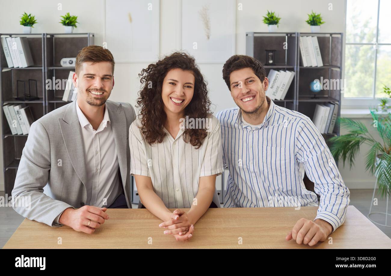 Happy young coworkers sitting at desk in office during informal meeting and looking at camera. Stock Photo