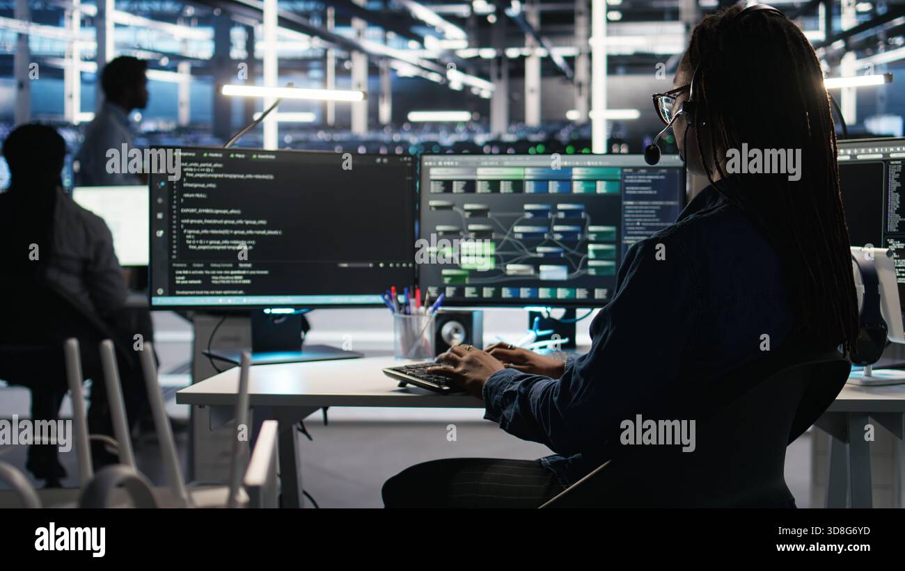 Data center employee uses audio headset and visual programming dashboard to troubleshoot client reported issues. Server farm technician looks at node tree interface, talking in microphone Stock Photo
