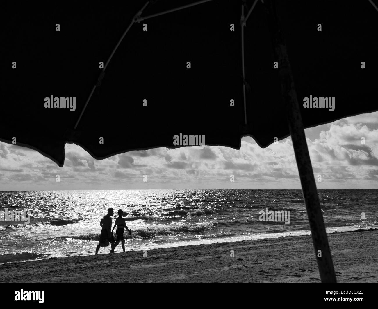 Silhouetted couple walking along a sunlit shoreline, framed beneath the shadow of a large beach umbrella in a dramatic black-and-white coastal scene. - Smartphone Captured Stock Image