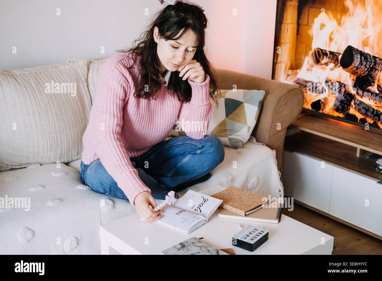 Young adult woman sits on sofa writing goals for year in notebook next to fireplace display. Goal setting, personal development, self reflection, cozy Stock Photo