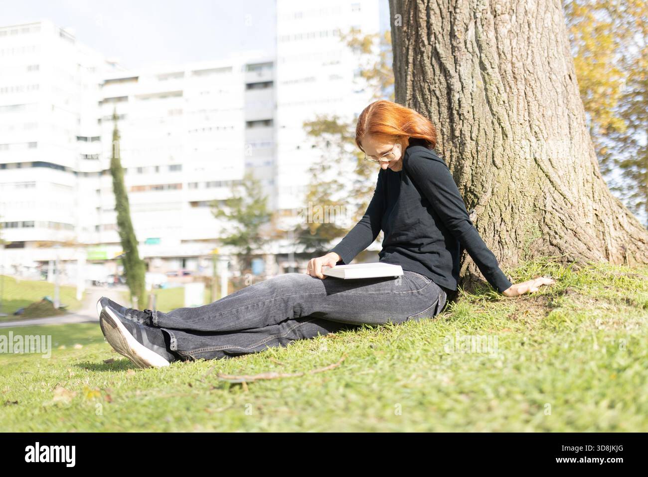 Young woman reading book leaning against tree in park Stock Photo