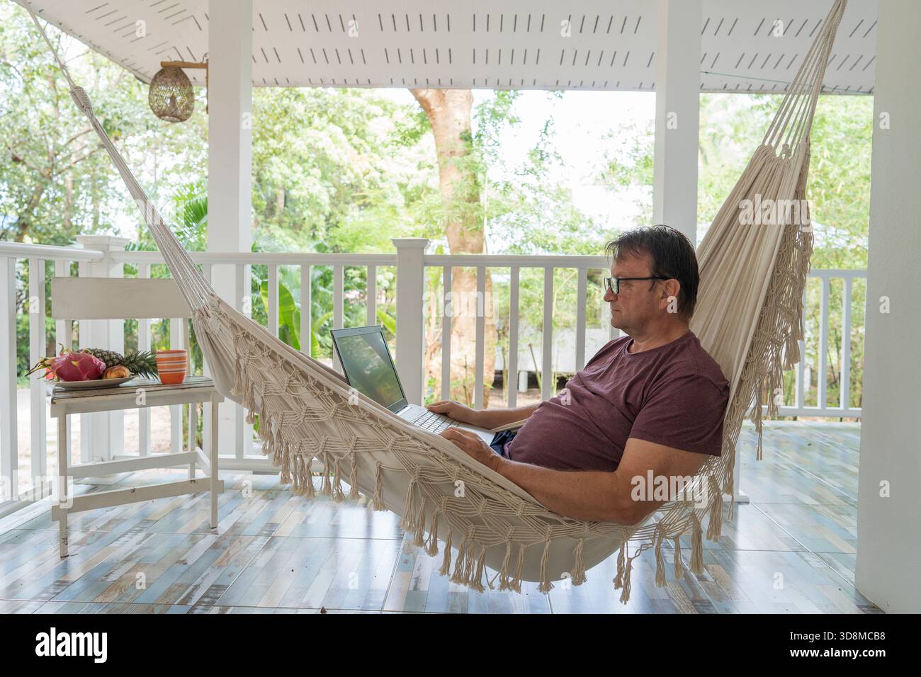 Grown man rests and working a laptop in a hammock on a terrace home near tropical garden in island Koh Phangan, Thailand, close up Stock Photo