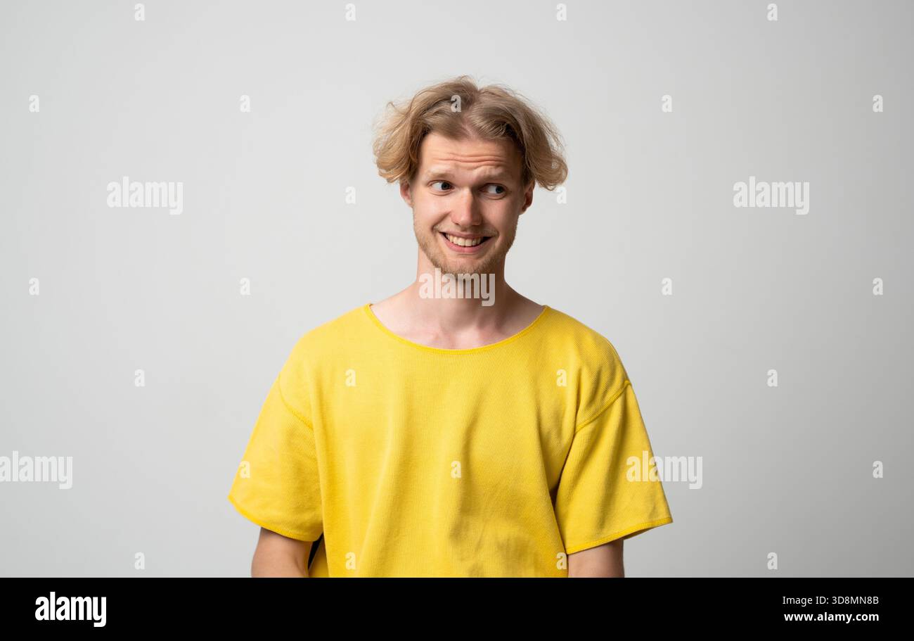 Smiling young man looking sideways with awkward amused expression while posing in neutral studio scene Stock Photo