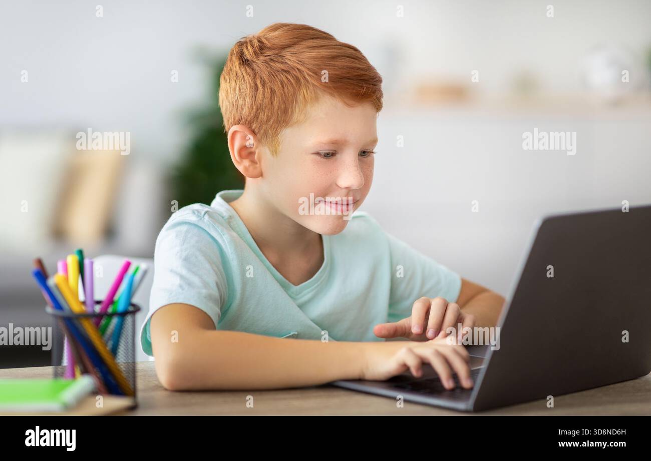 Joyful Ginger Boy Focused on Homework While Using Laptop at Modern Desk Stock Photo