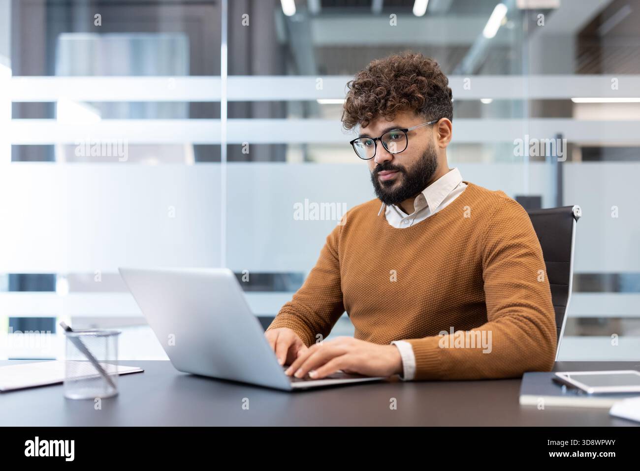 Young diverse businessman with glasses and beard working intently on a laptop at a contemporary desk in a bright, modern office, focusing on his tasks and demonstrating productivity Stock Photo