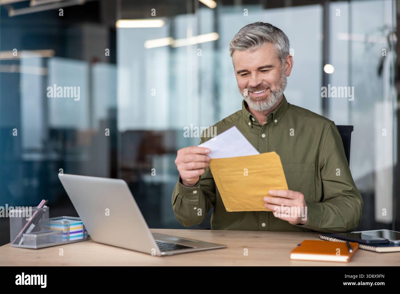 Mature cheerful businessman sitting at his office desk, happily opening a physical mail envelope with important documents, an old-school alternative to digital correspondence, while using a laptop Stock Photo