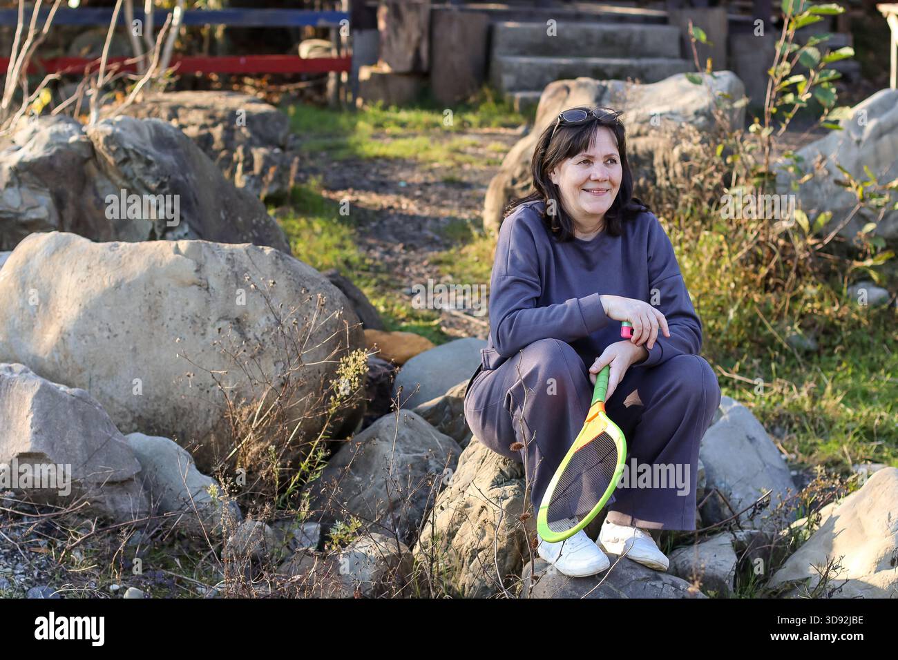 Mature woman in blue outfit sits on large stones, smiling while holding yellow and green badminton racket in grassy area with path. Stock Photo