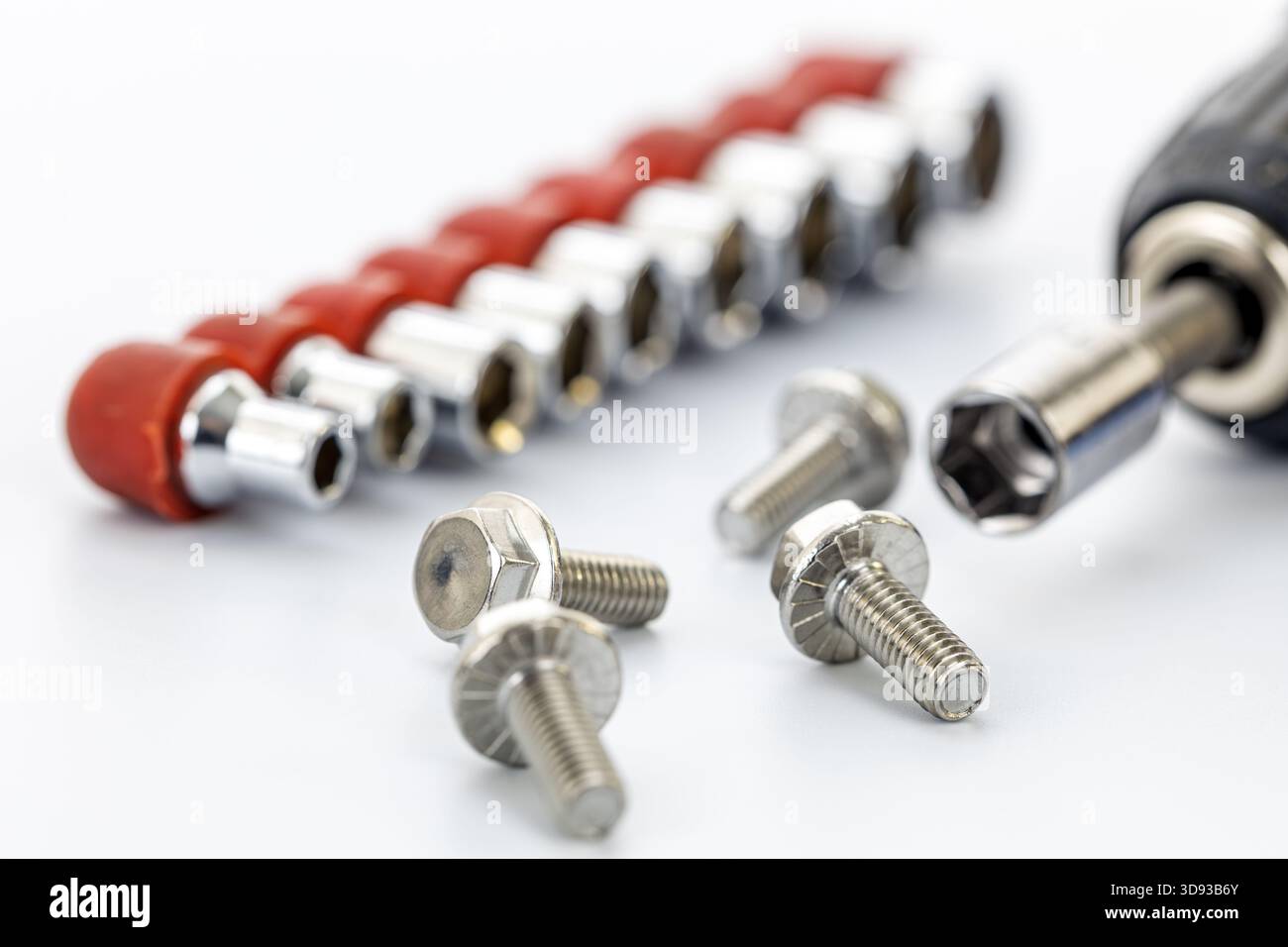 A close-up of five silver metal screws placed on a white surface with a socket wrench attachment and a row of various socket bits with red holders in Stock Photo