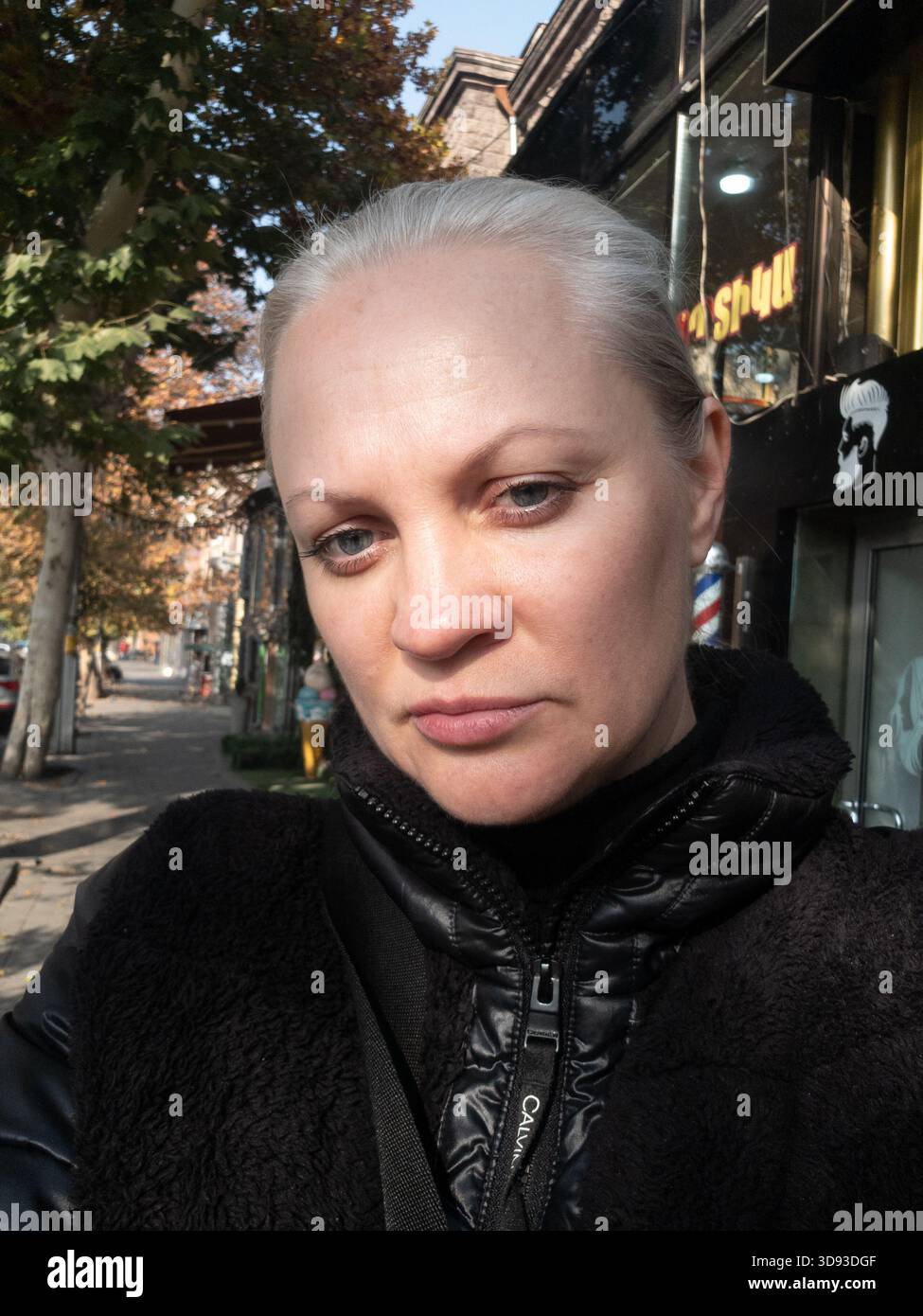A woman with gray hair is standing on a sidewalk in front of a building. She is wearing a black coat and a scarf Stock Photo