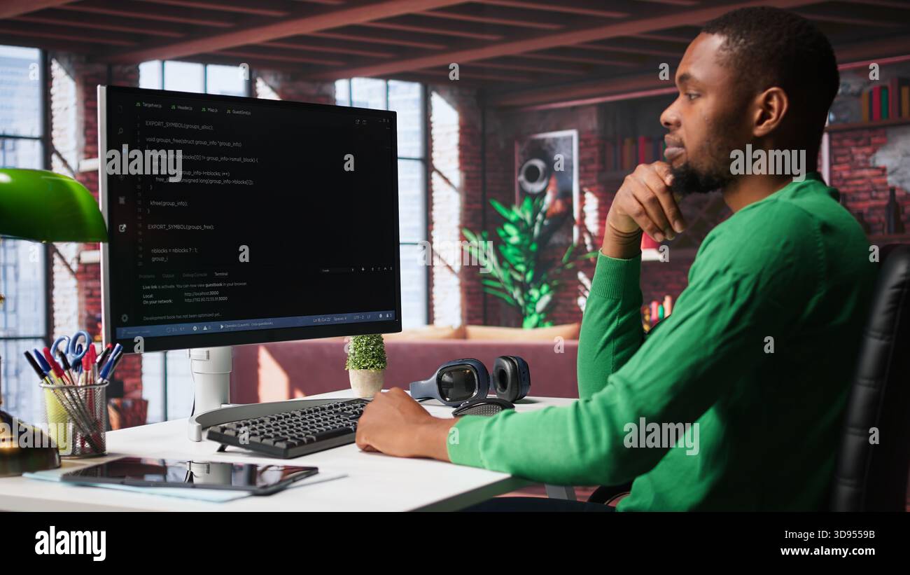 Black technician in home office looks at UI on computer monitor, typing and executing programming code. IT worker using coding application to write and test code in modern apartment. Camera B. Stock Photo