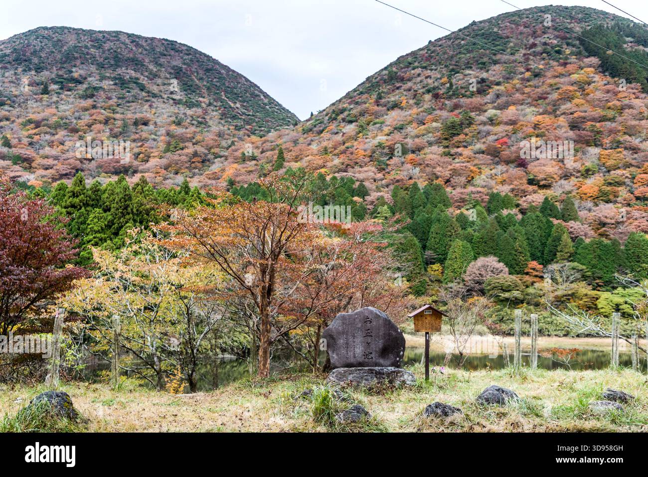 A commemorative stone bearing an inscription in Japanese calligraphy on the banks of a pond near the Amazake Chaya in Hakone, Kanagawa Prefecture, Japan. In the background are the autumn-coloured slopes of the Hakone Mountains; to the left is a wooden information sign Stock Photo
