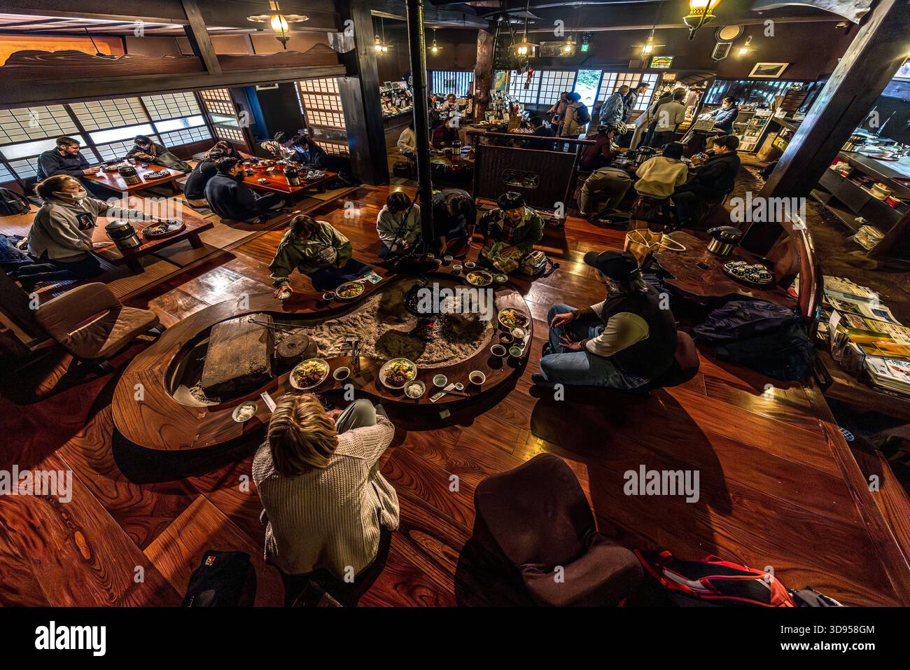 A bird’s-eye view of the bustling dining area of an irori restaurant in Hakone, Kanagawa Prefecture, Japan: guests sit on the wooden floor around a large circular irori (sunken hearth) with a cast-iron cauldron, arranging their bowls of food. The irori serves as a cooking area, a source of heat and the focal point for socialising; the combination of a wooden beamed ceiling, shoji windows and the smoke from the fire is typical of traditional mountain villages in the Japanese Alps Stock Photo