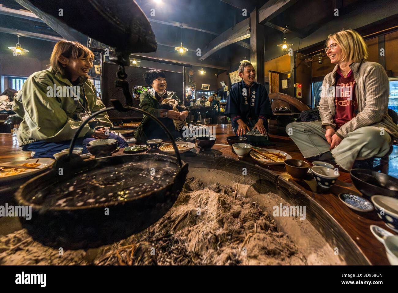Four people are sitting around an irori in a traditional restaurant in Hakone, Kanagawa Prefecture, Japan: a member of staff in blue uniform and three guests, including two non-Japanese women, are chatting whilst eating together. In front of them, ceramic bowls containing Japanese dishes are arranged around the smouldering hearth. Irori restaurants offer guests an introduction to Japan’s traditional hearth cuisine Stock Photo