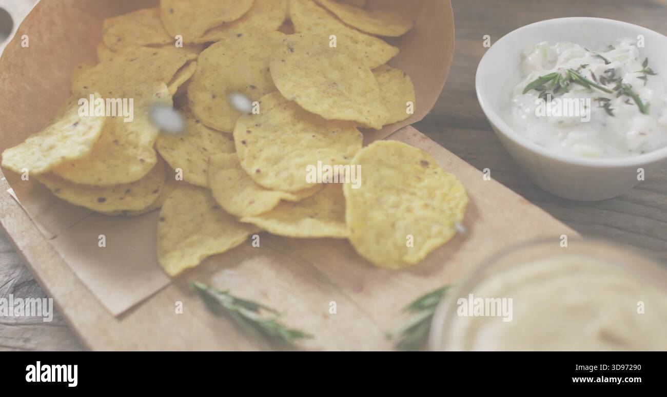 Spilling corn tortilla chips from paper cone onto wooden board on rustic tabletop, with herb dip Stock Photo
