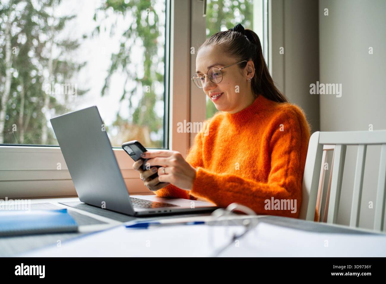 Work from home with phone and laptop. Woman with computer on kitchen table. Entrepreneur making business content with smartphone. Happy freelancer. Stock Photo