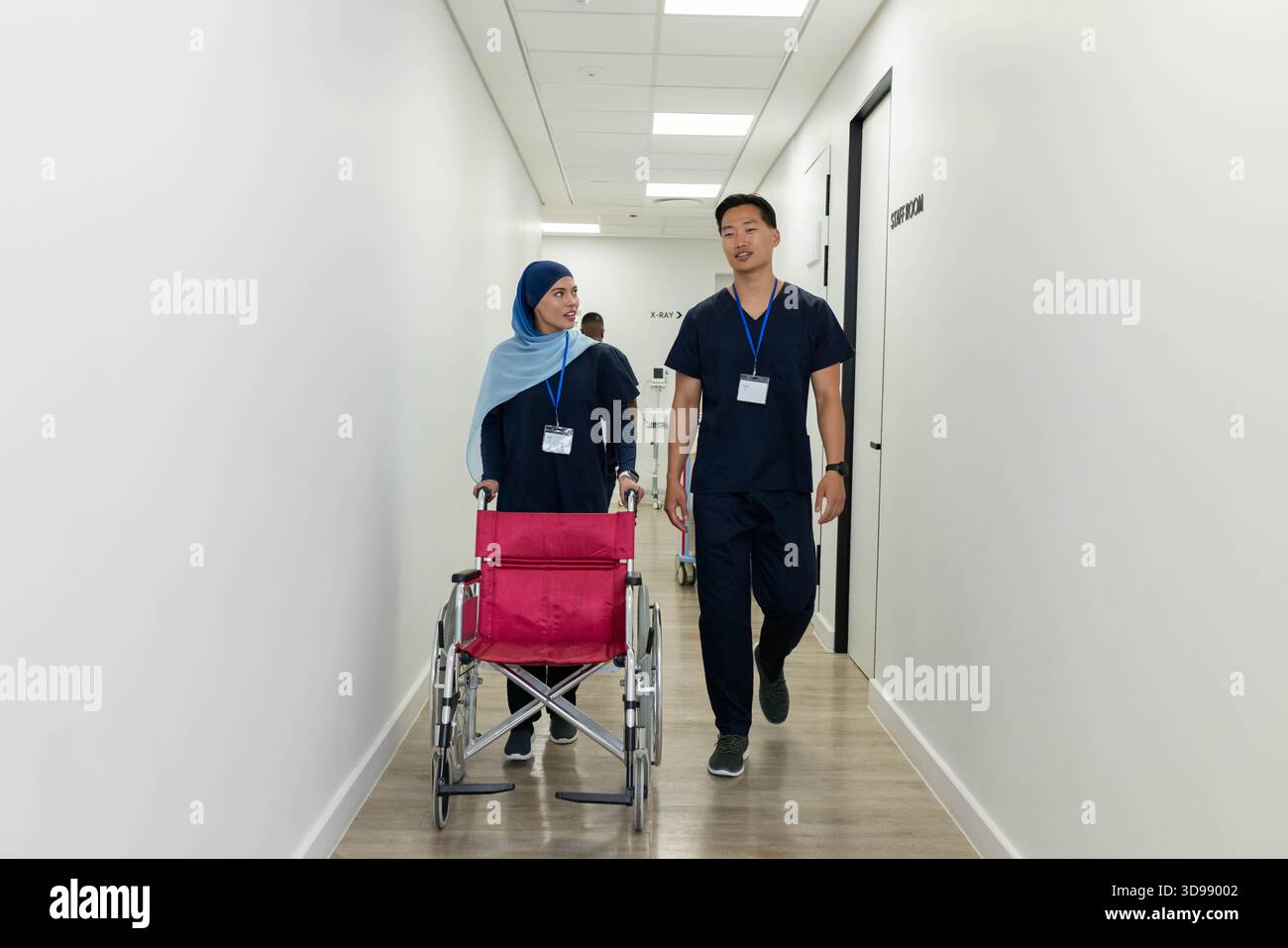 Diverse coworkers in scrubs walking hospital hall, pushing magenta-seat wheelchair with ID badges Stock Photo
