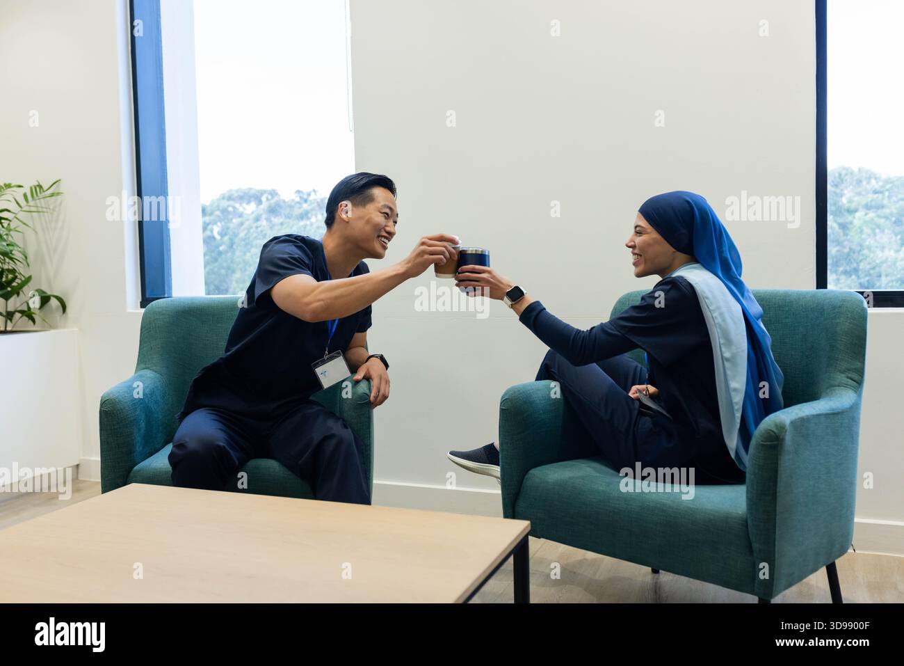 Diverse coworkers in navy scrubs clinking travel mugs while sitting in staff break room Stock Photo