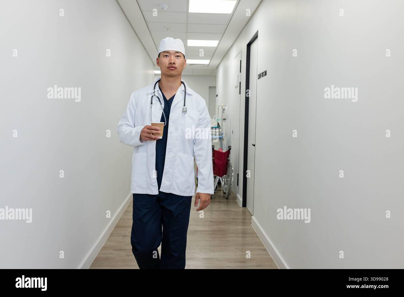 Asian male doctor walking hospital hall navy scrubs, white lab coat, holding paper cup, stethoscope Stock Photo