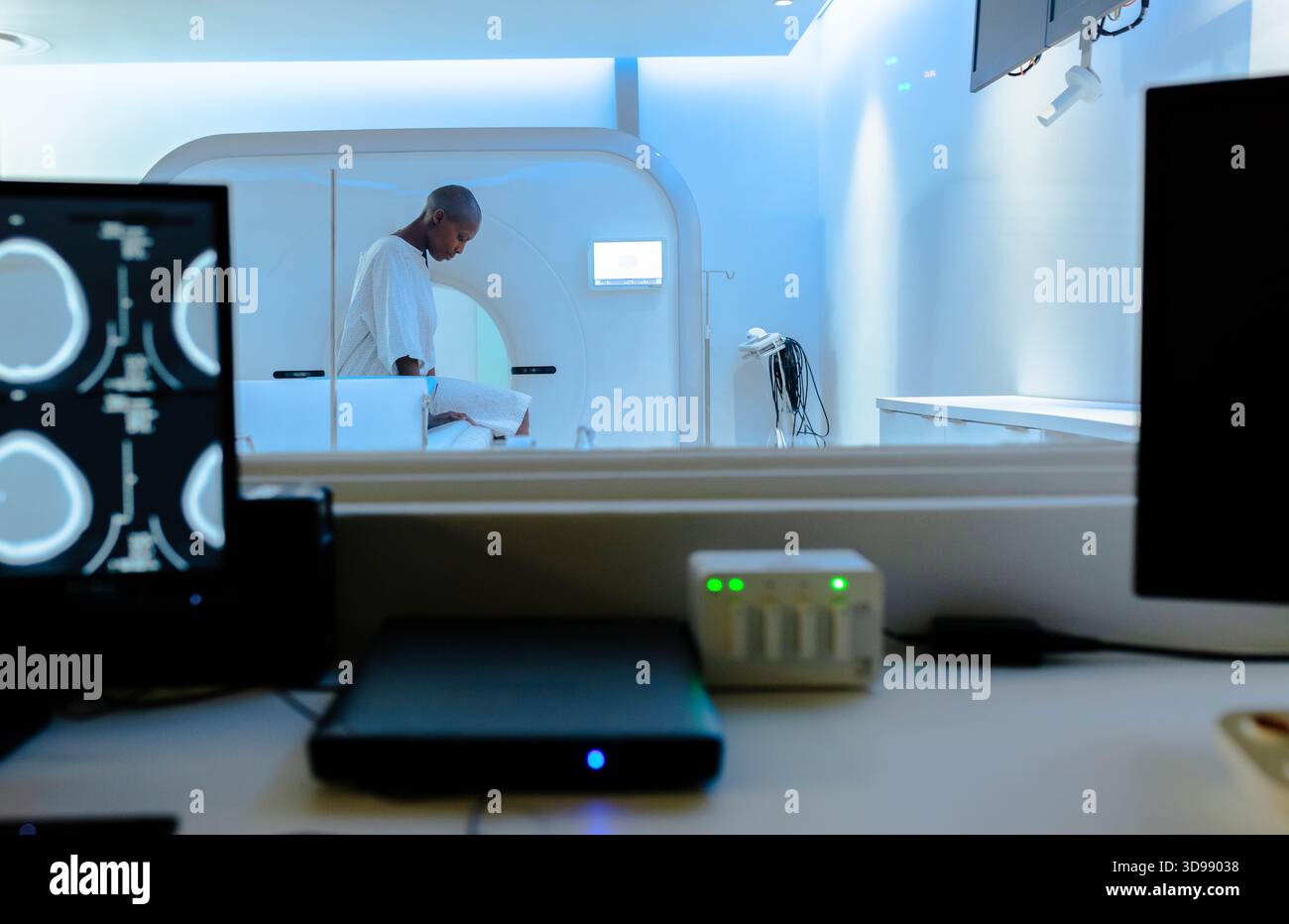 Adult African woman patient standing in exam gown beside MRI gantry table behind control monitors Stock Photo