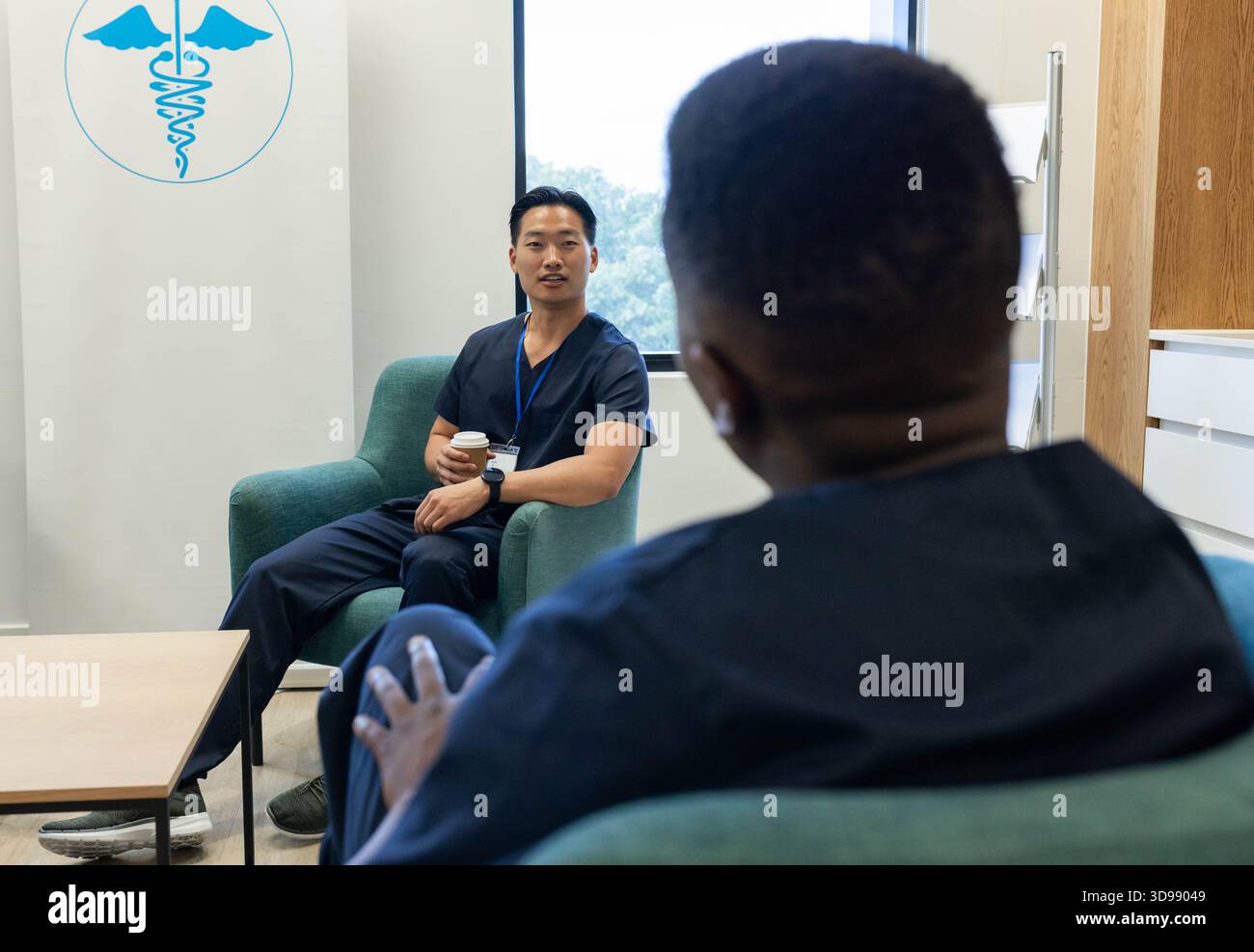 Diverse male colleagues in navy scrubs sitting in staff lounge discussing case with coffee lanyard Stock Photo