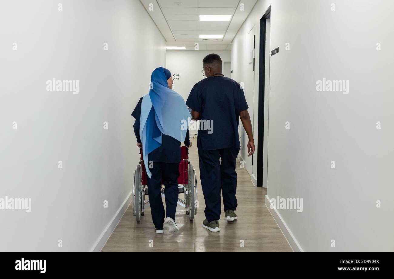African American nurses walking hospital hall pushing red wheelchair wearing navy scrubs and hijab Stock Photo