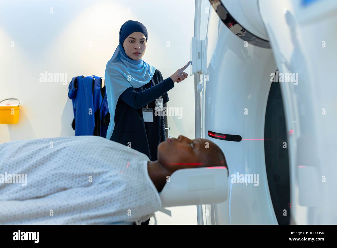 CT technologist in blue hijab adjusting CT gantry while male patient lying under red laser lines Stock Photo