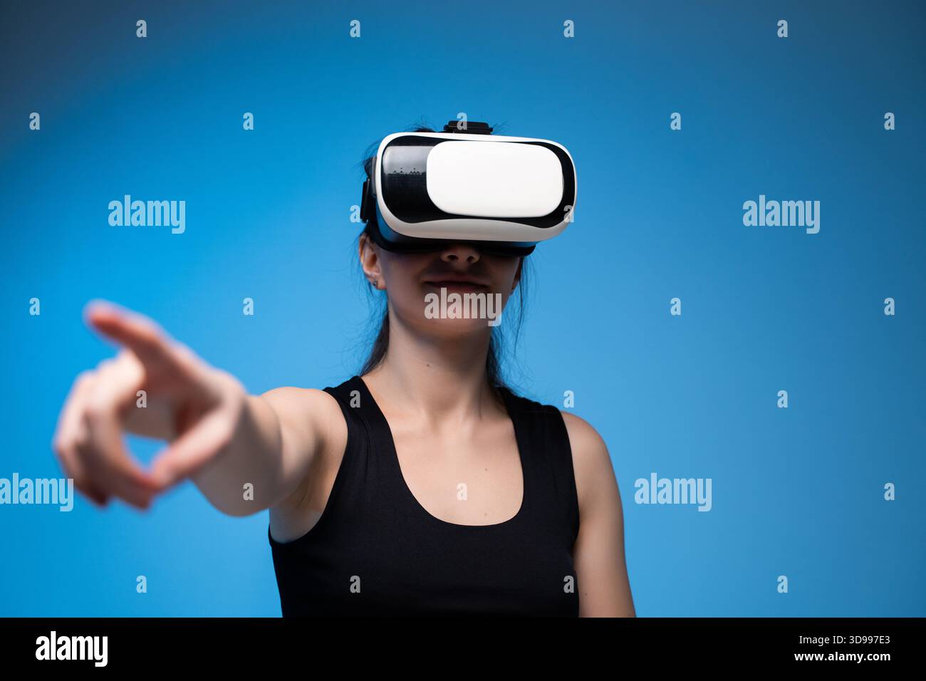 Woman wearing VR headset pointing forward while engaging with virtual interface in digital blue studio Stock Photo
