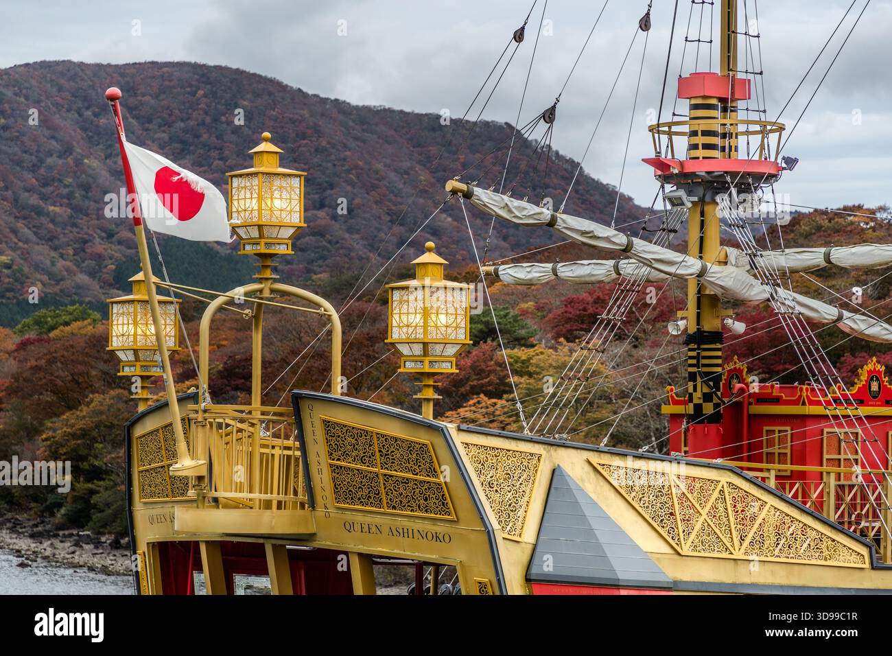 The bow and upper deck of the excursion boat ‘Queen Ashinoko’ on Lake Ashi in Hakone, Kanagawa Prefecture, Japan. The gold-decorated ship, flying the Japanese flag, is moored against a mountainside covered in autumnal deciduous woodland; the Queen Ashinoko operates as part of the Hakone Sightseeing Cruise on the crater lake in the Hakone Mountains Stock Photo