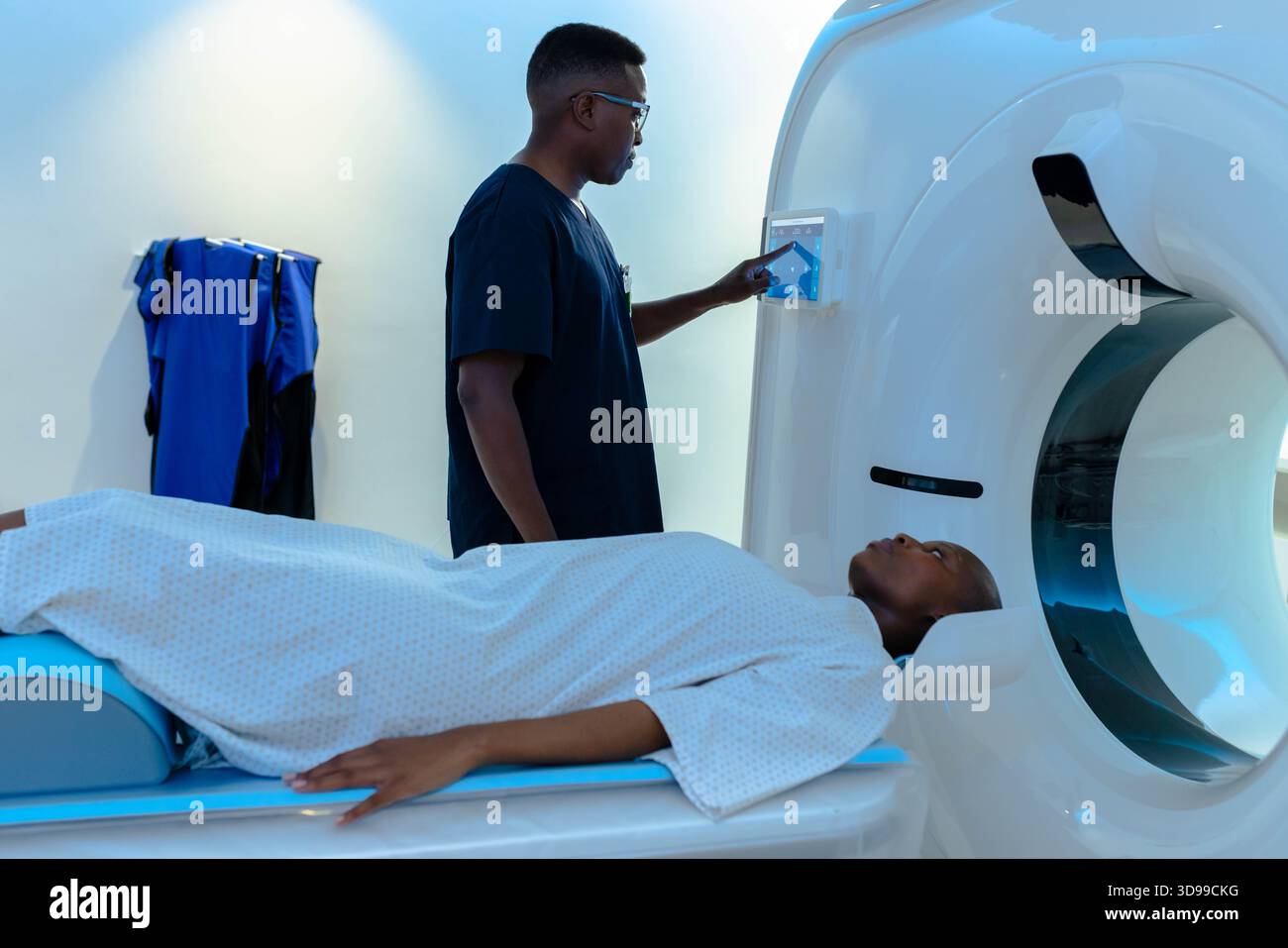 Patient sliding into scanner gantry in radiology room, tech adjusting panel, blue aprons hanging Stock Photo