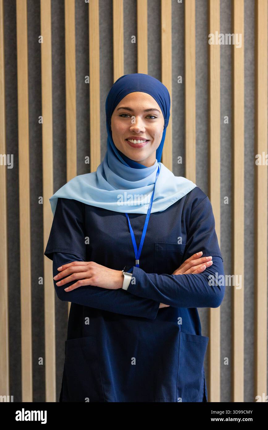 Woman posing arms crossed smiling in navy scrubs, headscarf, ID badge, smartwatch at clinic Stock Photo