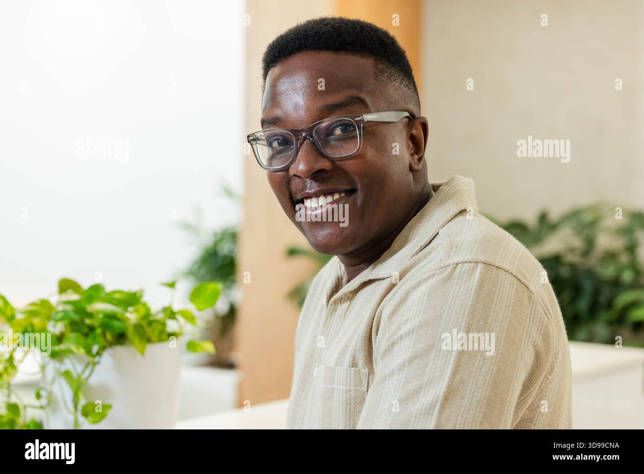 Adult African American man smiling in lounge in tan shirt and clear glasses beside white planter Stock Photo