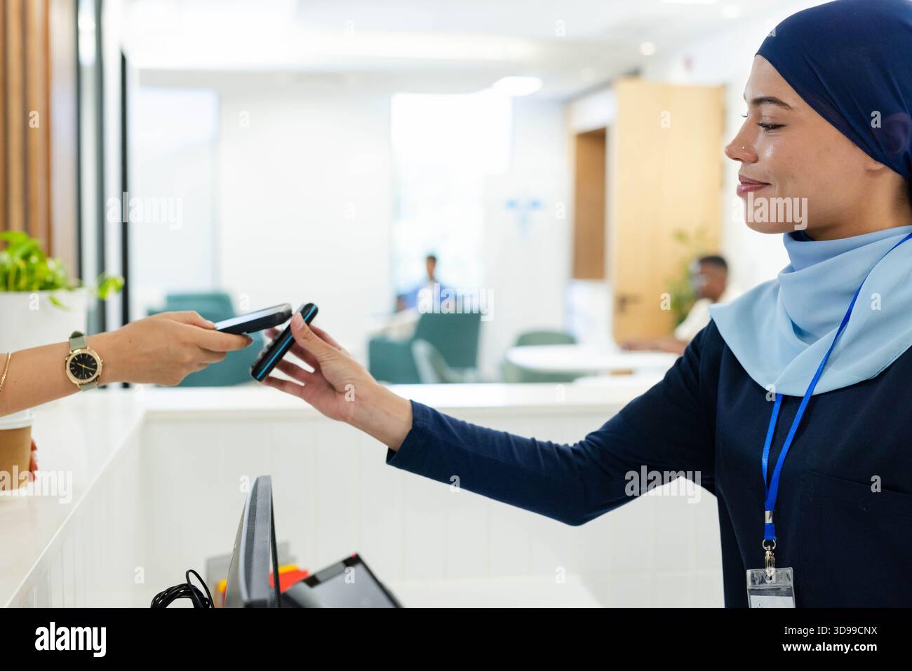 Receptionist extending card reader toward customer holding smartphone at bright reception counter Stock Photo