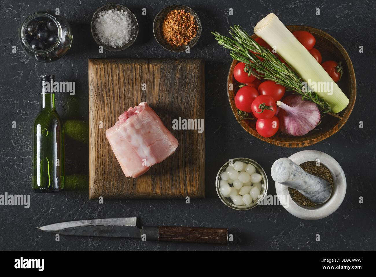 Fresh ingredients are beautifully arranged on a wooden board, including a piece of raw rabbit saddle meat, ripe tomatoes, garlic, leeks, olives, and h Stock Photo