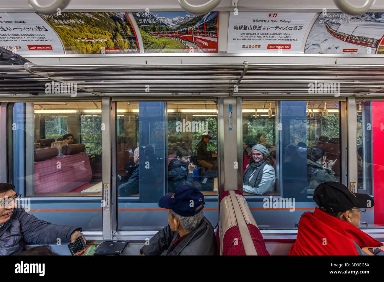 Passengers inside a carriage on the Hakone Tozan Railway (箱根登山鉄道), the rack railway in the mountainous Hakone region of Kanagawa Prefecture. A poster on the ceiling advertises the partnership between the Hakone Tozan Railway and the Rhaetian Railway in Switzerland (Rhaetian Railway); both lines connect their respective mountain regions via steeply graded routes. The wooded slopes of the Hakone mountain range can be seen through the carriage windows. Hakone, Kanagawa Prefecture, Japan Stock Photo